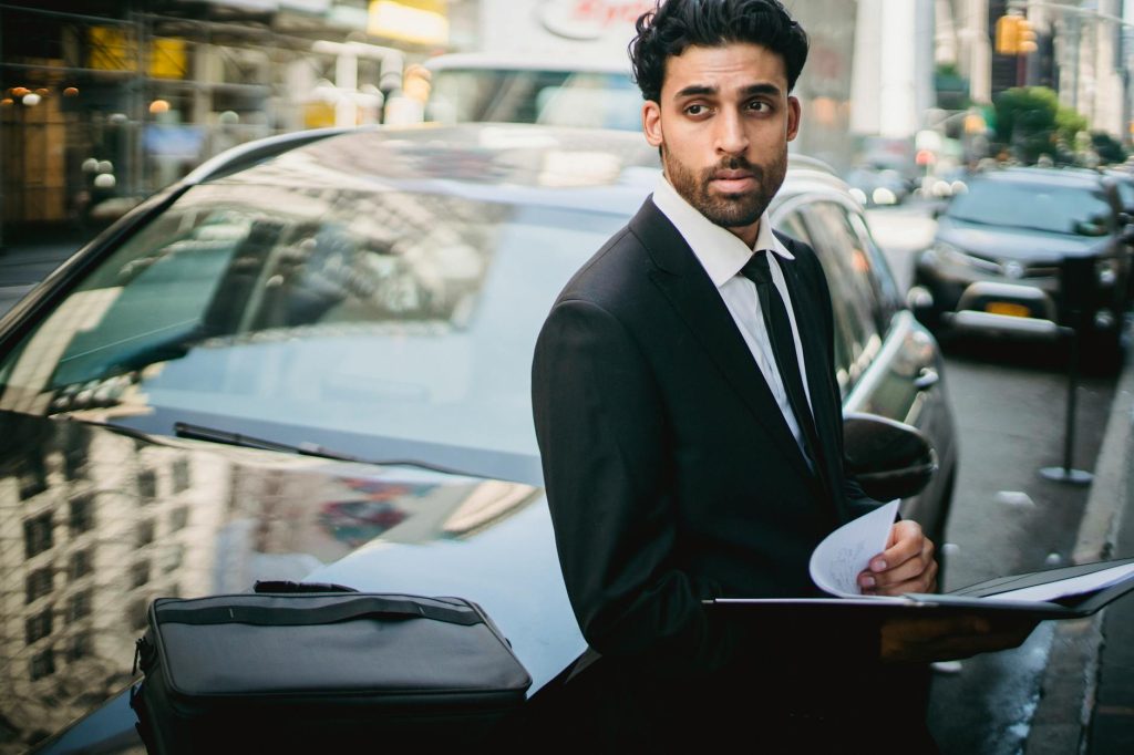 Stylish businessman in a suit leans on a car in a bustling city street.