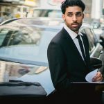 Stylish businessman in a suit leans on a car in a bustling city street.