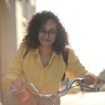 From below cheerful young curly woman in glasses with backpack smiling at camera while riding bicycle along street in sunny day