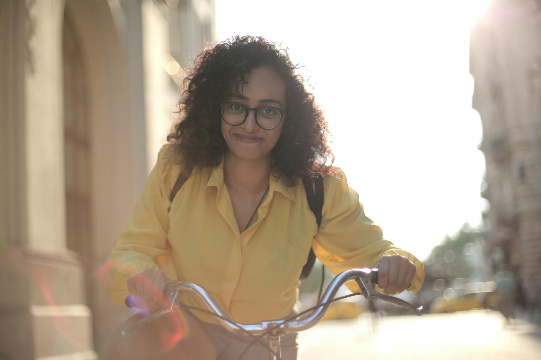 From below cheerful young curly woman in glasses with backpack smiling at camera while riding bicycle along street in sunny day