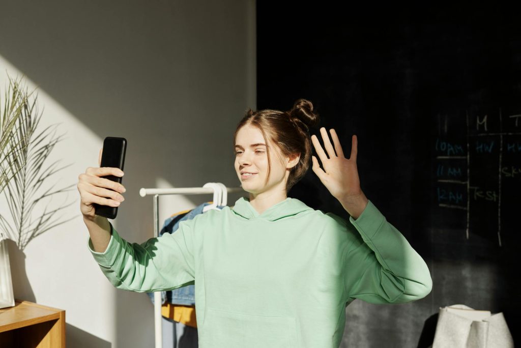 Young woman using smartphone for a video call indoors, waving and smiling in natural sunlight.