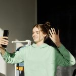 Young woman using smartphone for a video call indoors, waving and smiling in natural sunlight.