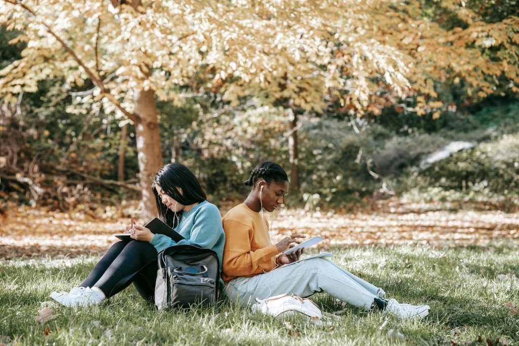 Two young women study with gadgets in a vibrant fall park setting, highlighting diverse learning environments.