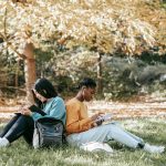 Two young women study with gadgets in a vibrant fall park setting, highlighting diverse learning environments.