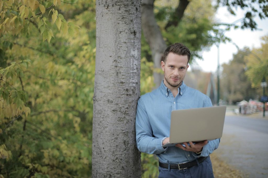 Confident young male freelancer in casual shirt and jeans leaning on tree and using laptop while spending time in park in city and looking at camera