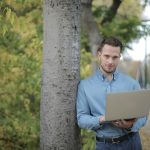 Confident young male freelancer in casual shirt and jeans leaning on tree and using laptop while spending time in park in city and looking at camera