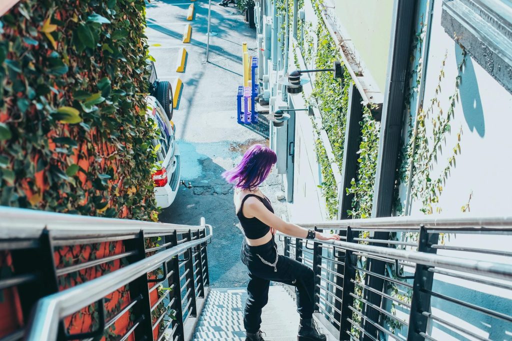 A woman with vibrant purple hair descends a staircase in an urban setting, exuding a modern and fashionable vibe.