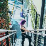 A woman with vibrant purple hair descends a staircase in an urban setting, exuding a modern and fashionable vibe.
