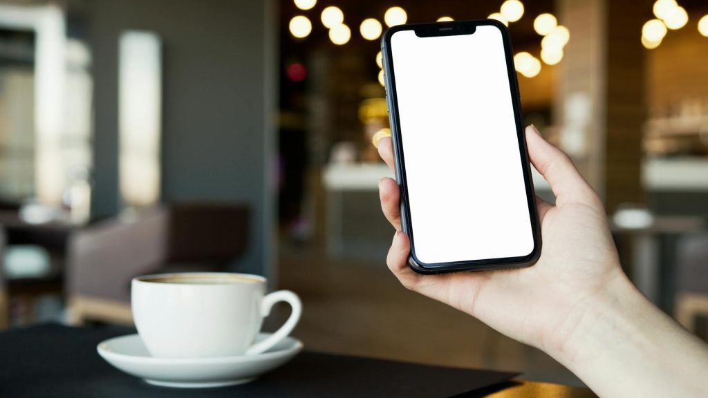 Hand holding smartphone with blank screen and coffee cup in cozy cafe setting.