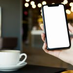Hand holding smartphone with blank screen and coffee cup in cozy cafe setting.