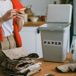 A person sorting recyclable materials into a labeled paper bin in a kitchen setting.
