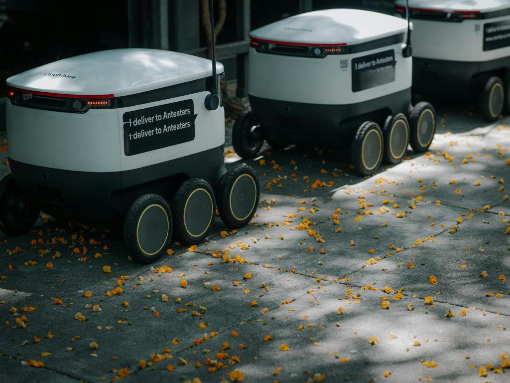 Futuristic delivery robots navigating a leaf-strewn sidewalk, showcasing innovation in modern urban logistics.