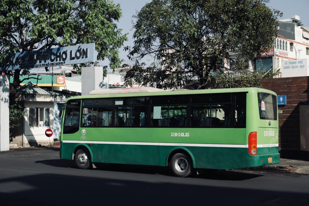 Green public bus parked on a street in Ho Chi Minh City, Vietnam, under a tree on a sunny day.