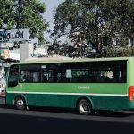 Green public bus parked on a street in Ho Chi Minh City, Vietnam, under a tree on a sunny day.