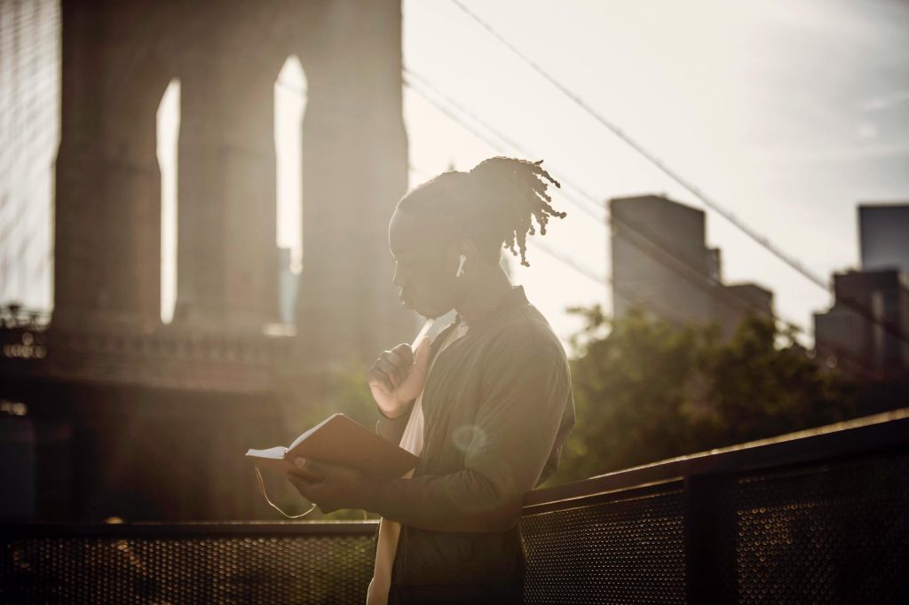 Side view of pensive young black man with braided hairstyle reading book and listening to music with wireless earphones while standing on city promenade against bridge