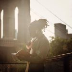 Side view of pensive young black man with braided hairstyle reading book and listening to music with wireless earphones while standing on city promenade against bridge